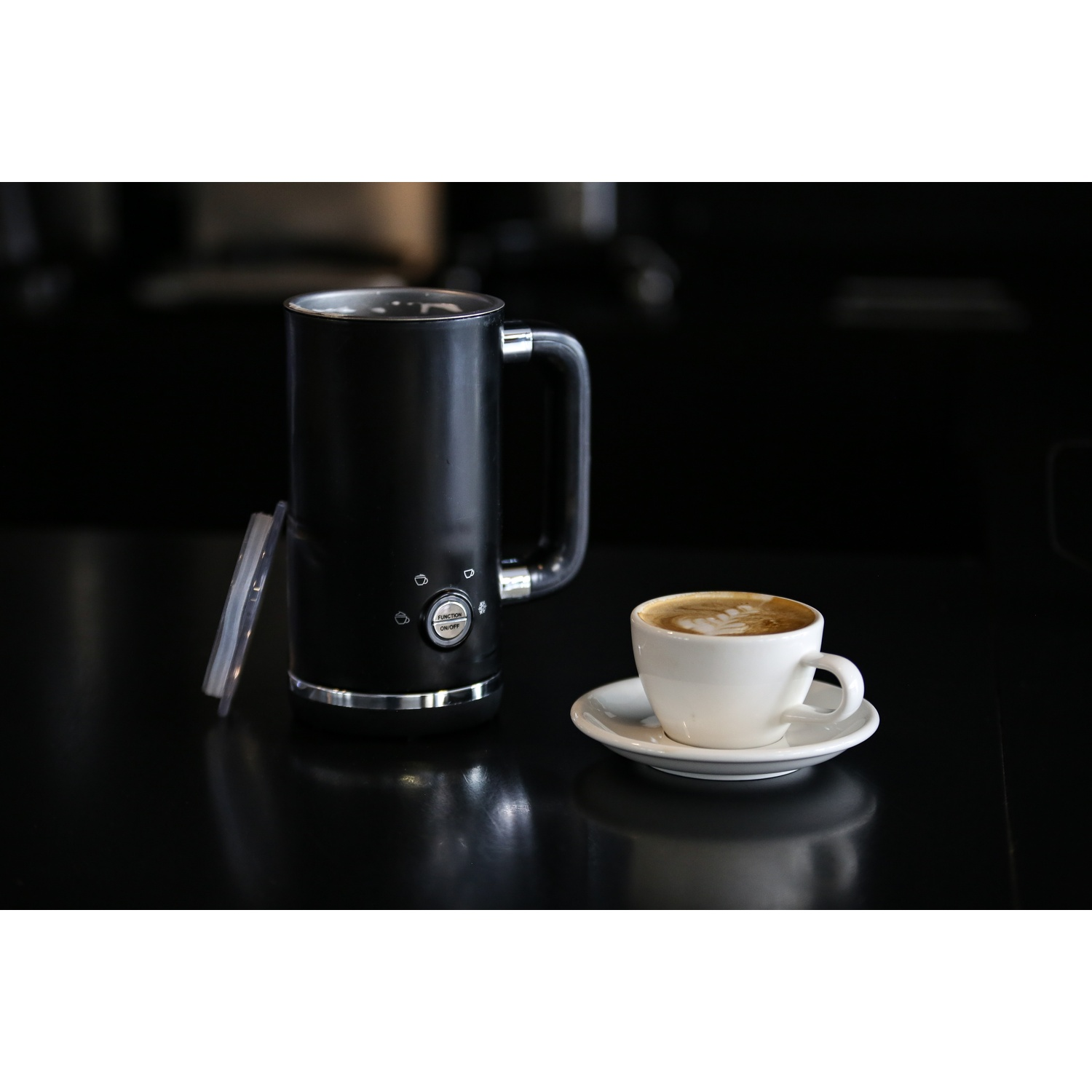 Un montalatte nero con manico, una tazza bianca di caffè con latte art e un piattino su una superficie nera.