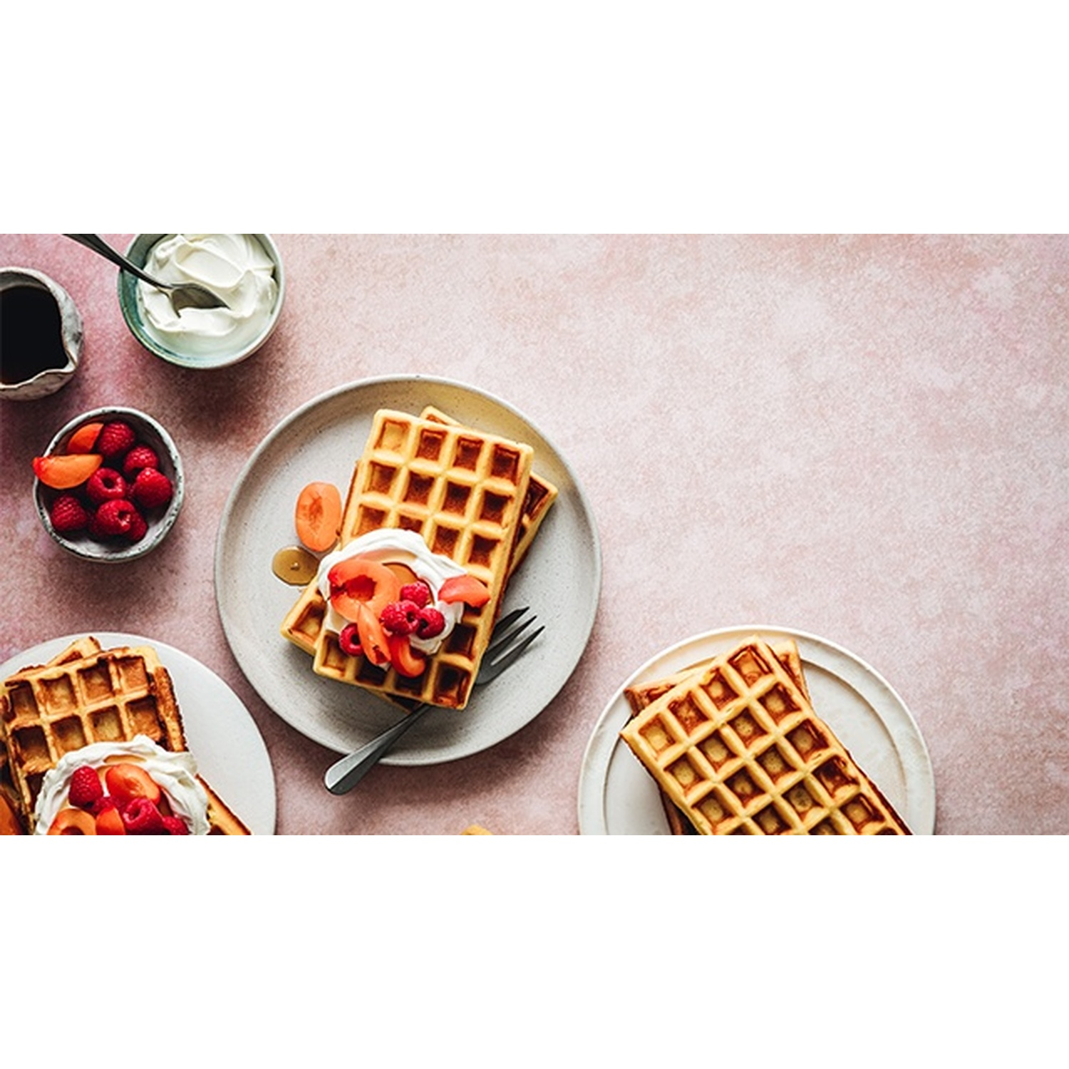 Waffle con frutta e panna nei piatti. Ciotole di frutta e panna. Sfondo rosa. Sciroppo e forchetta visibili.
