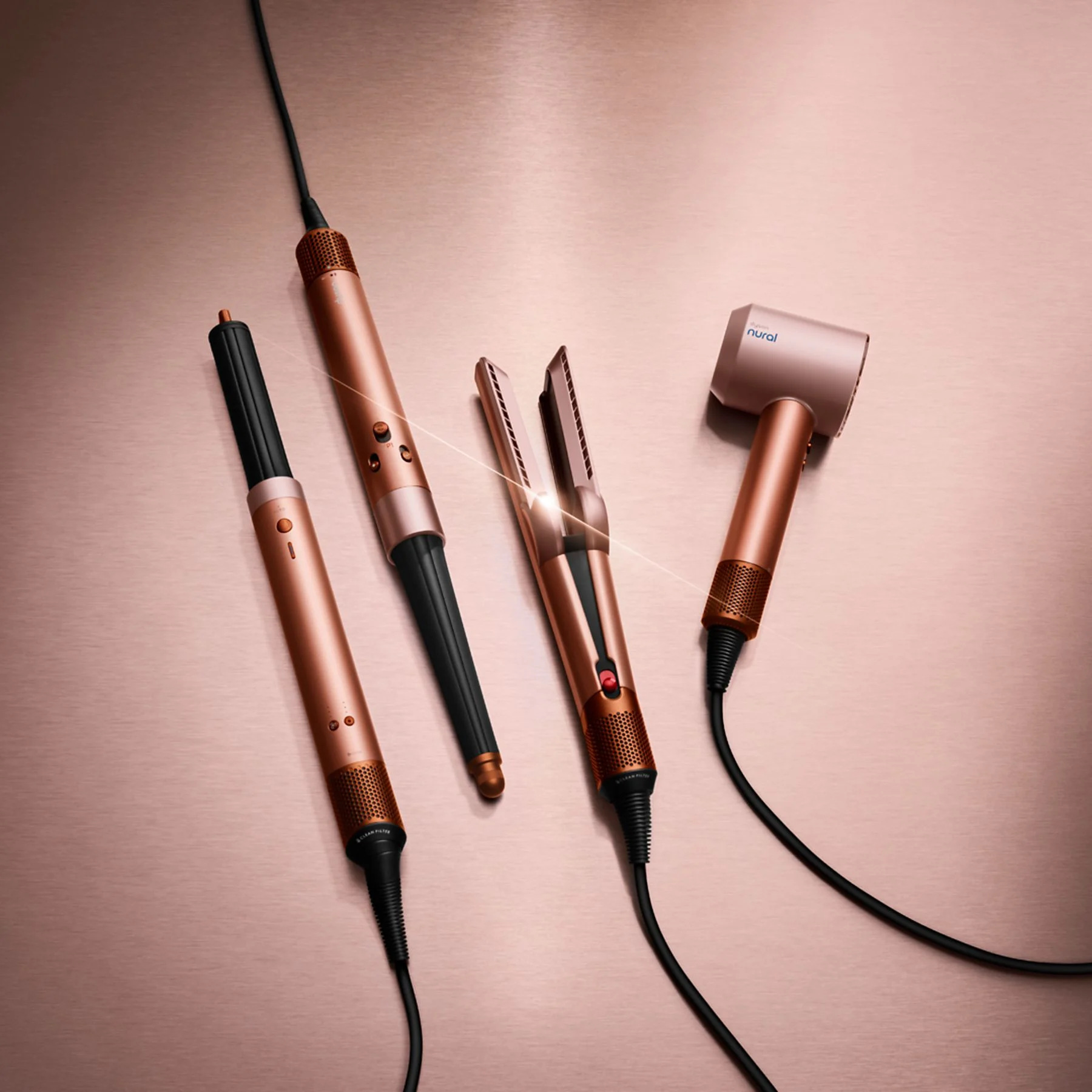 Cztery miedziane narzędzia do stylizacji włosów na odblaskowej, różowo-złotej powierzchni. Zawiera suszarkę, prostownicę i dwa stylizatory.