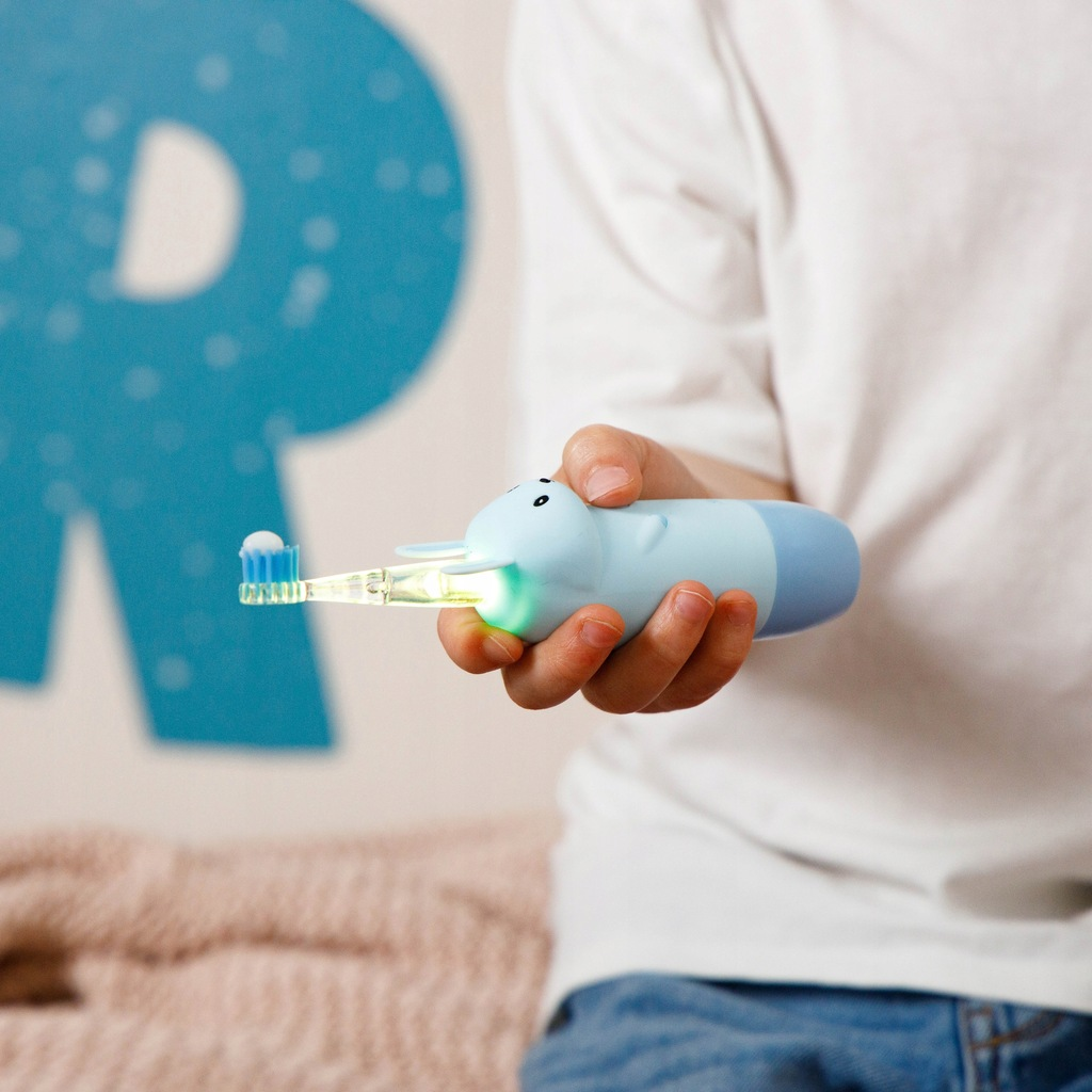Dziecko trzyma niebieską szczoteczkę do zębów z motywem królika. Szczoteczka ma pastę do zębów. Tło: ściana z niebieską literą R.