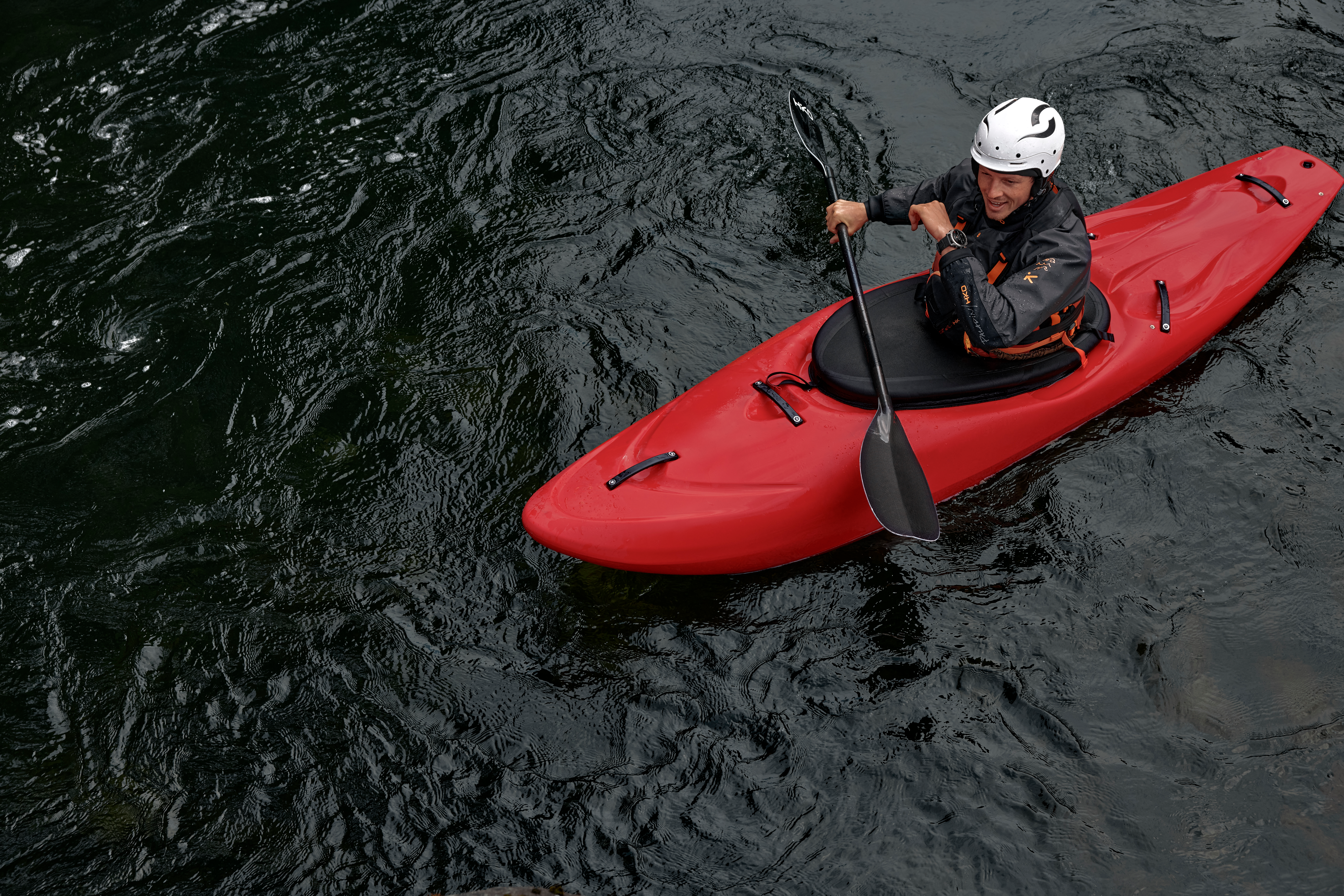 Una persona in un kayak rosso pagaia su acqua scura. La persona indossa un casco e una giacca scura. L'acqua è scura e increspata.