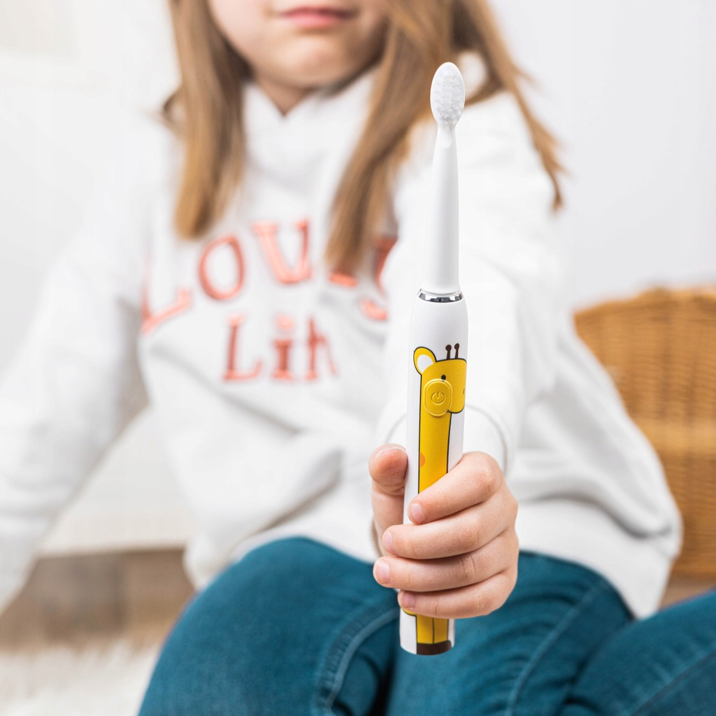 Dziewczyna trzyma szczoteczkę do zębów z motywem żyrafy. Szczoteczka jest biało-żółta. Dziewczyna ma na sobie białą bluzę z kapturem.