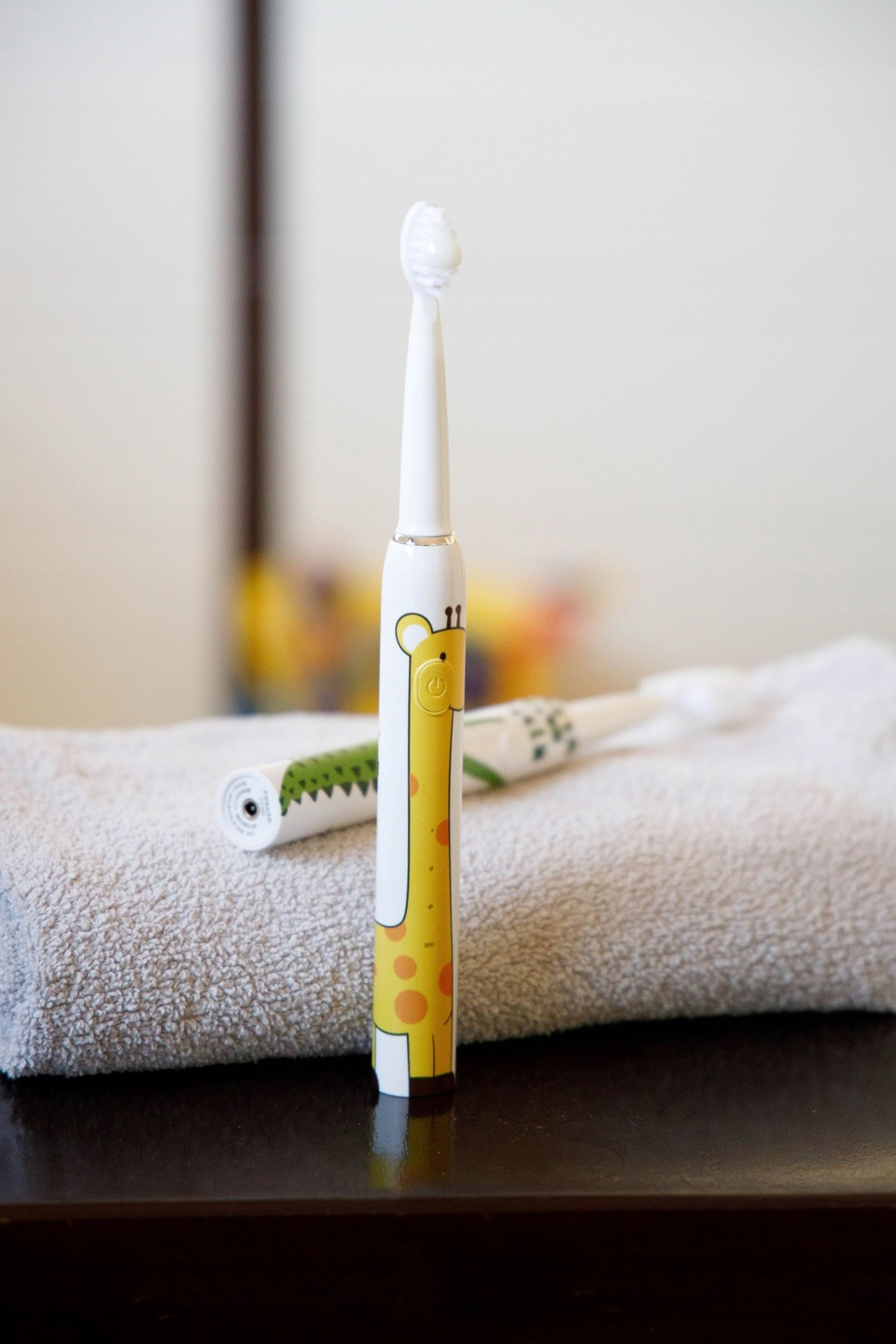 Szczoteczka do zębów dla dzieci z motywem żyrafy stoi na ręczniku, w tle kolejna szczoteczka. Szczoteczka jest biała i żółta.