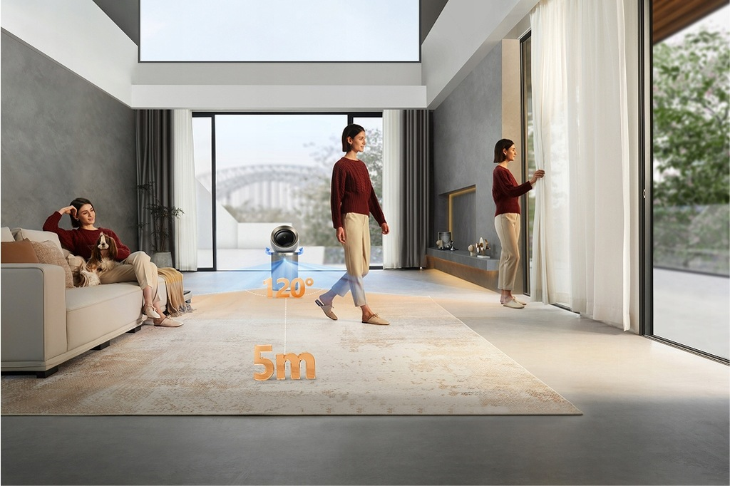 Kobieta spaceruje w nowoczesnym salonie. Inna siedzi na sofie z psem. Urządzenie stoi na podłodze, a po prawej stronie znajduje się okno.