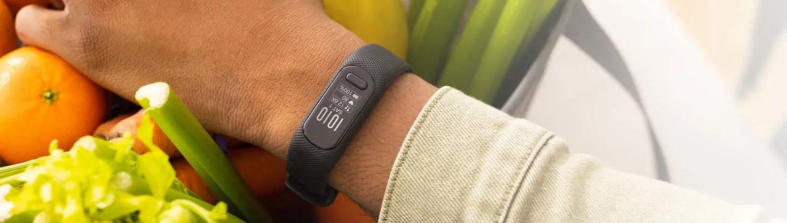Un polso con un fitness tracker nero che tiene verdure. Arance e sedano sono visibili in primo piano.