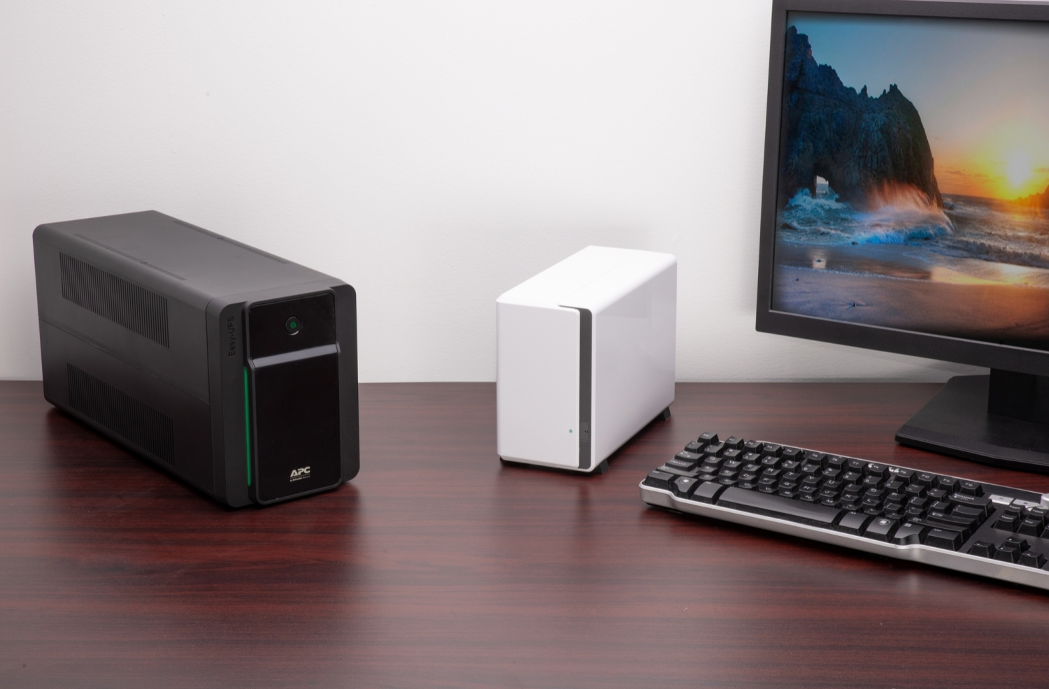 Biurko z czarnym zasilaczem UPS, białym NAS, monitorem z zachodem słońca i klawiaturą.