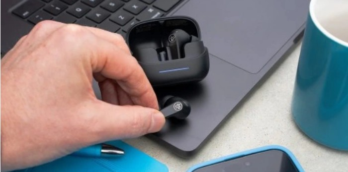 Ręka trzyma czarną słuchawkę. Etui i druga słuchawka leżą na laptopie. Niebieski kubek i telefon są w tle.