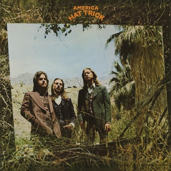 Albumcover von Americas 'Hat Trick'. Drei Männer in Anzügen stehen vor einer Wüstenlandschaft. Der Text lautet 'America Hat Trick'.