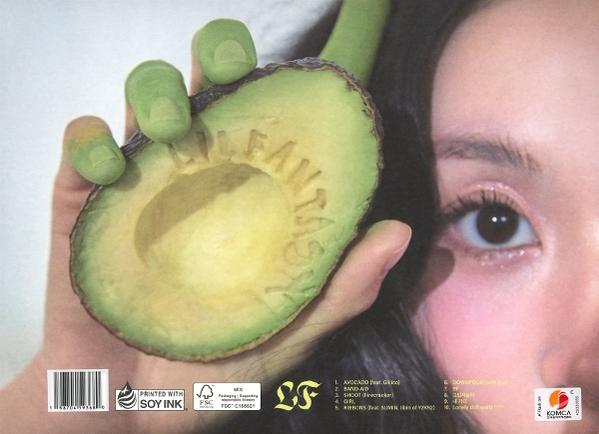 Eine Hand, die eine geschnittene Avocado hält, in die 'LITTLE FANTASY' geschnitzt ist. Auf der rechten Seite des Bildes ist ein Frauenauge zu sehen.
