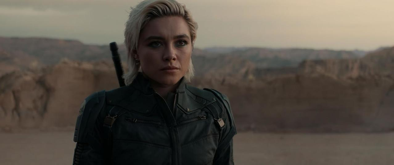 Eine Frau mit kurzen blonden Haaren steht in einer Wüstenlandschaft. Sie trägt eine dunkle Jacke und schaut in die Kamera.