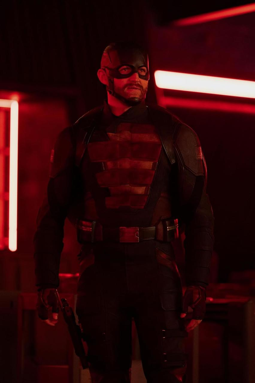 Ein Mann in schwarzer Uniform und Maske steht in einem rot beleuchteten Raum. Er hat eine Waffe in der Hand. Der Mann ist im Fokus.