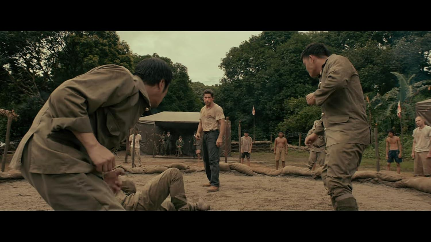 Männer in Militäruniformen und Freizeitkleidung befinden sich auf einem Feld. Ein Mann liegt am Boden. Andere schauen zu. Bäume und Zelte sind im Hintergrund.
