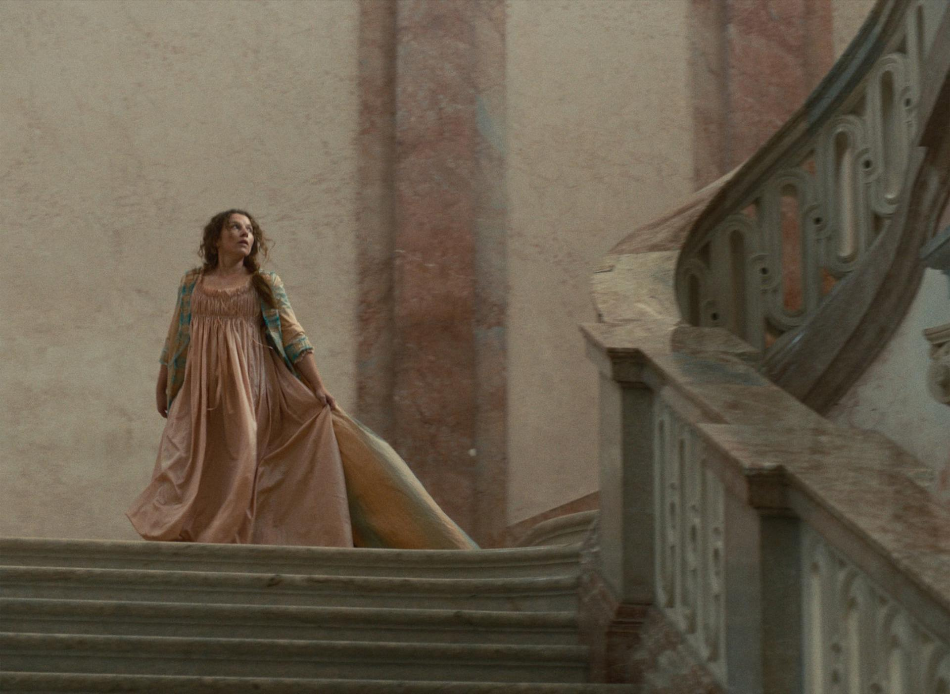 Eine Frau in einem pfirsichfarbenen Kleid und einer gemusterten Jacke steht auf einer großen Treppe und blickt nach oben. Im Hintergrund sind eine hohe Wand und Säulen zu sehen.