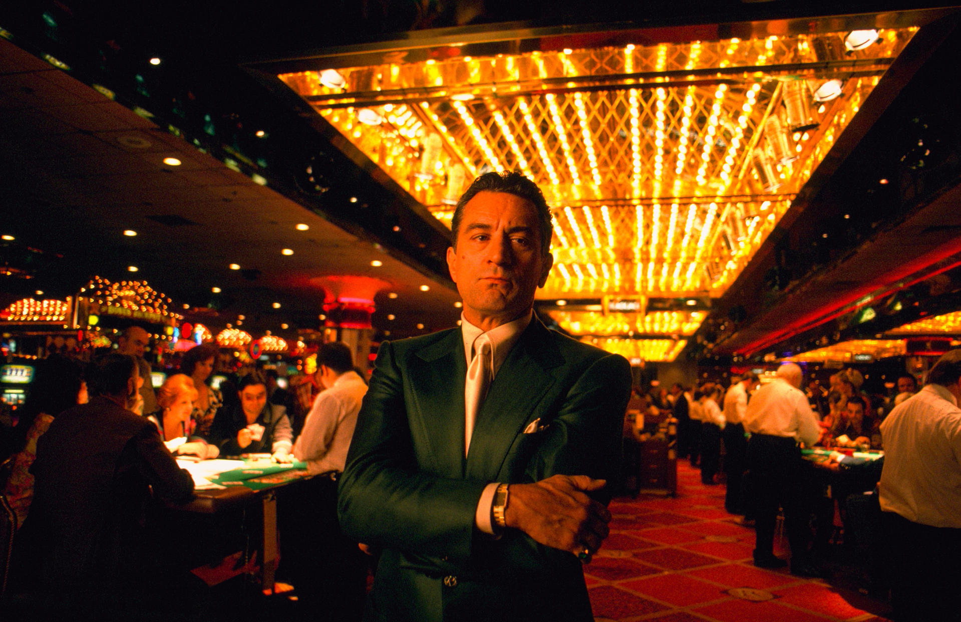 Mann im grünen Anzug steht im Casino. Helle Lichter oben. Im Hintergrund spielen Leute Karten. Roter Teppich und Tische.