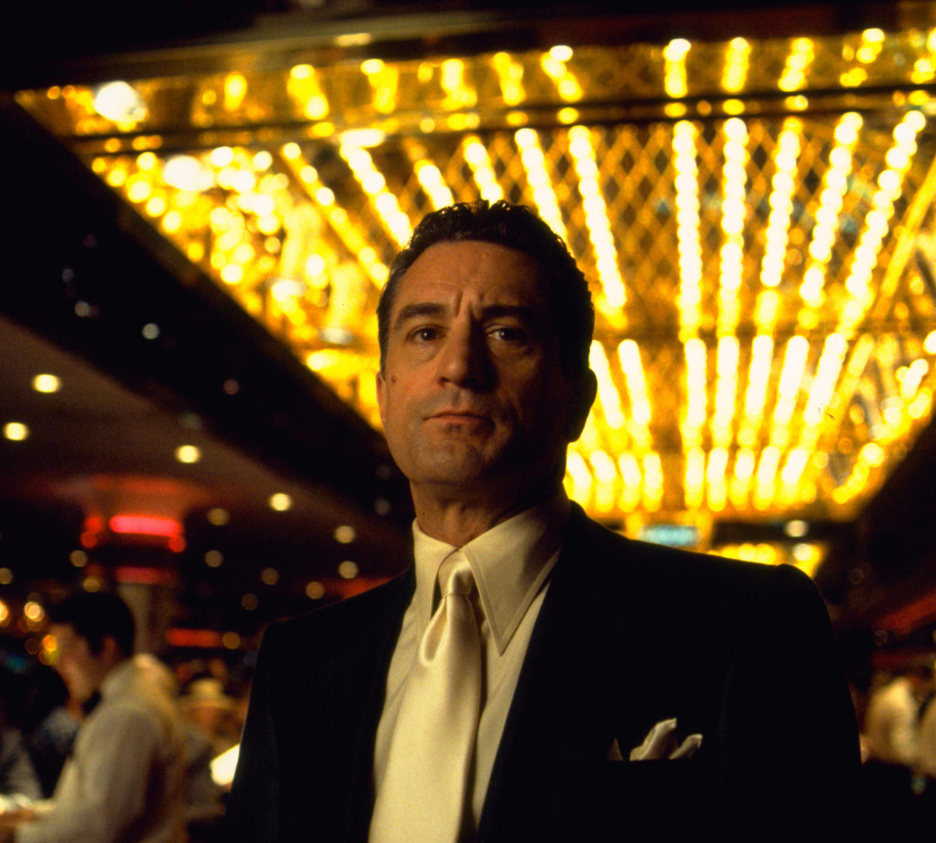 Mann im Anzug, weiße Krawatte, von goldenen Lichtern beleuchtet. Casino-Kulisse. Dunkler Hintergrund mit Menschen.
