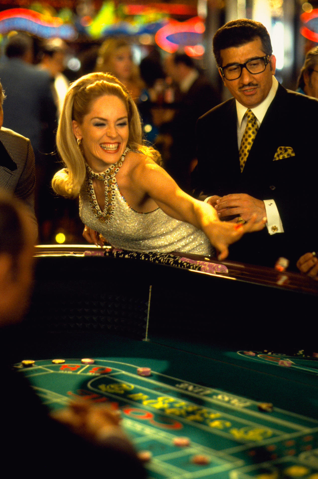 Eine Frau lächelt und greift nach einem Casinotisch. Ein Mann im Anzug steht neben ihr. Chips und Roulette-Tisch sind zu sehen.