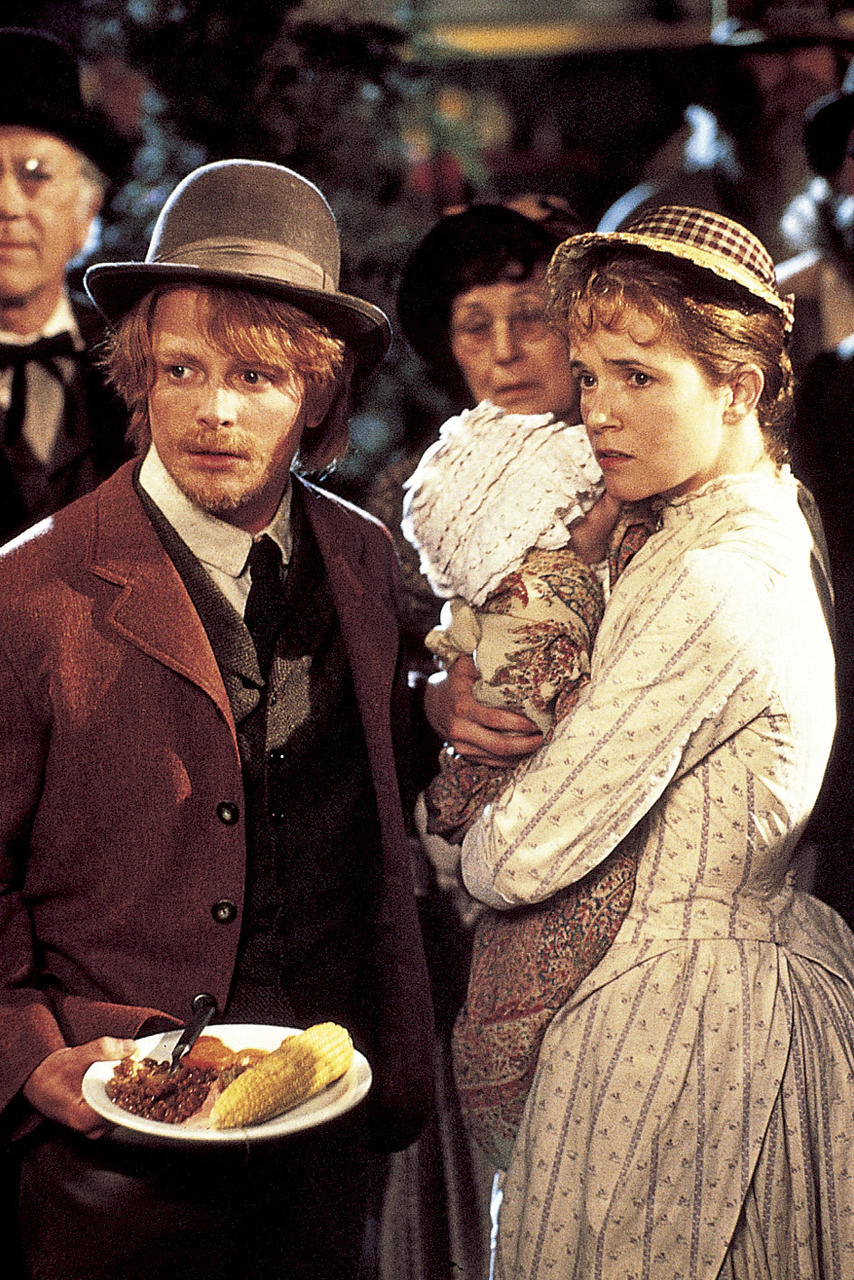 Ein Mann mit Hut hält Essen. Eine Frau hält ein Baby. Andere Leute sind im Hintergrund. Die Szene scheint im Freien zu sein.