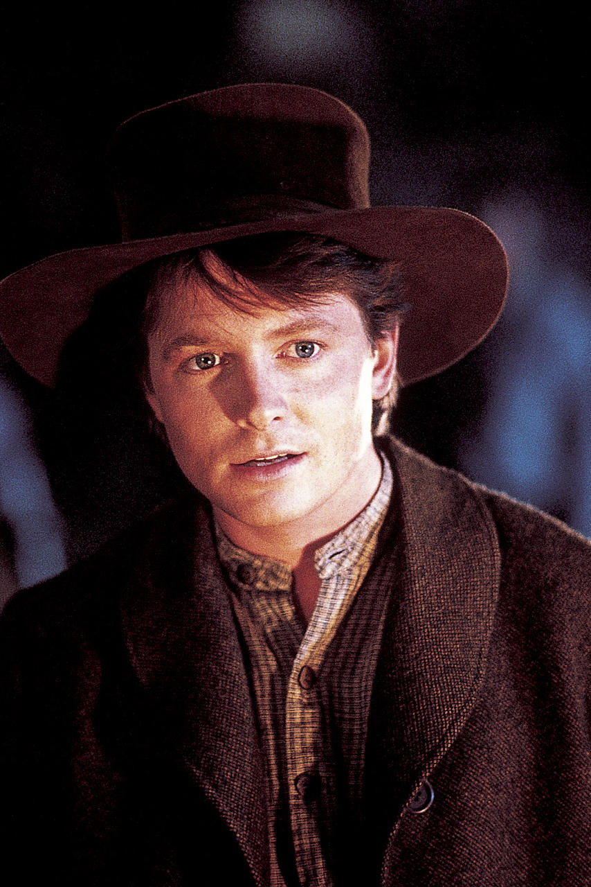 Mann mit braunem Hut und Mantel, der in die Kamera schaut. Der Hintergrund ist dunkel. Hemd sichtbar. Der Mann ist hellhäutig mit blauen Augen.