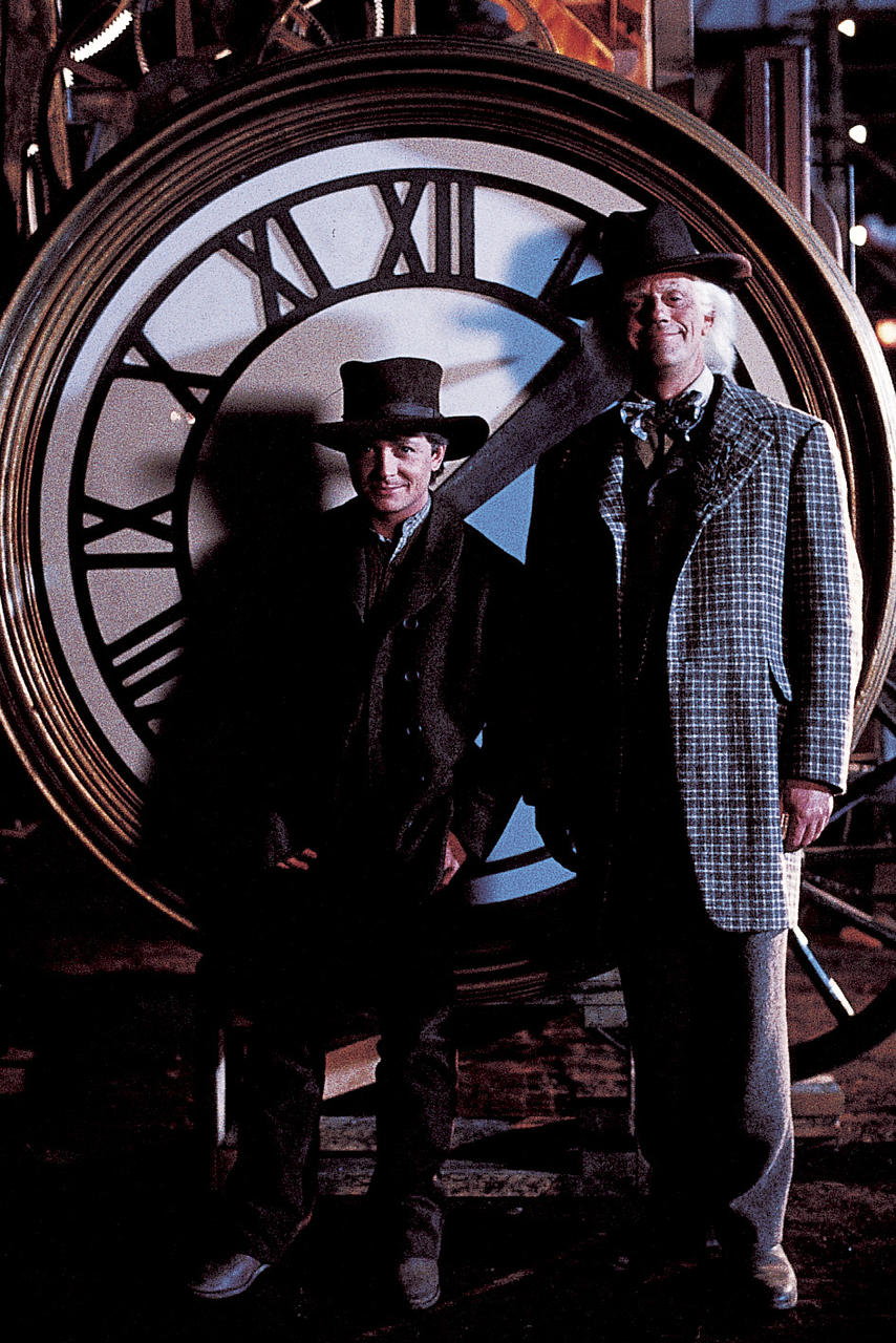 Zwei Männer mit Hüten stehen vor einem großen Zifferblatt. Einer trägt einen karierten Mantel. Der Hintergrund ist dunkel.