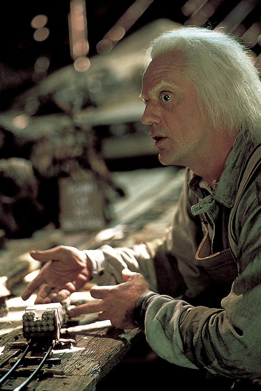 Ein älterer Mann mit weißem Haar deutet auf eine Miniatur-Eisenbahnstrecke. Er trägt eine grüne Jacke und sieht überrascht aus. Ein kleiner Zug steht auf den Gleisen.