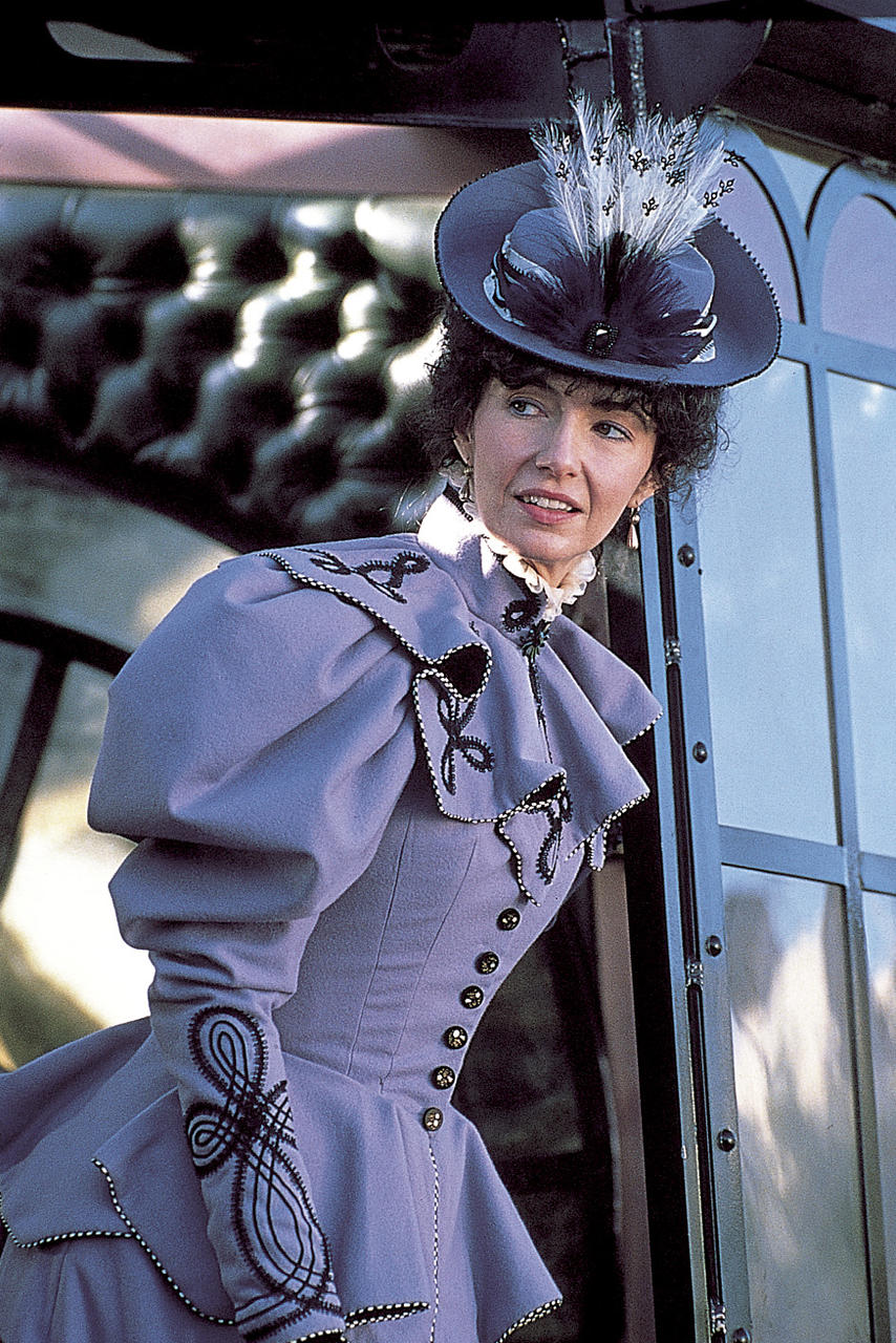 Frau in lila Kleid und Hut mit Federn steht an einem Fenster. Das Kleid hat aufwendige Verzierungen und Knöpfe.