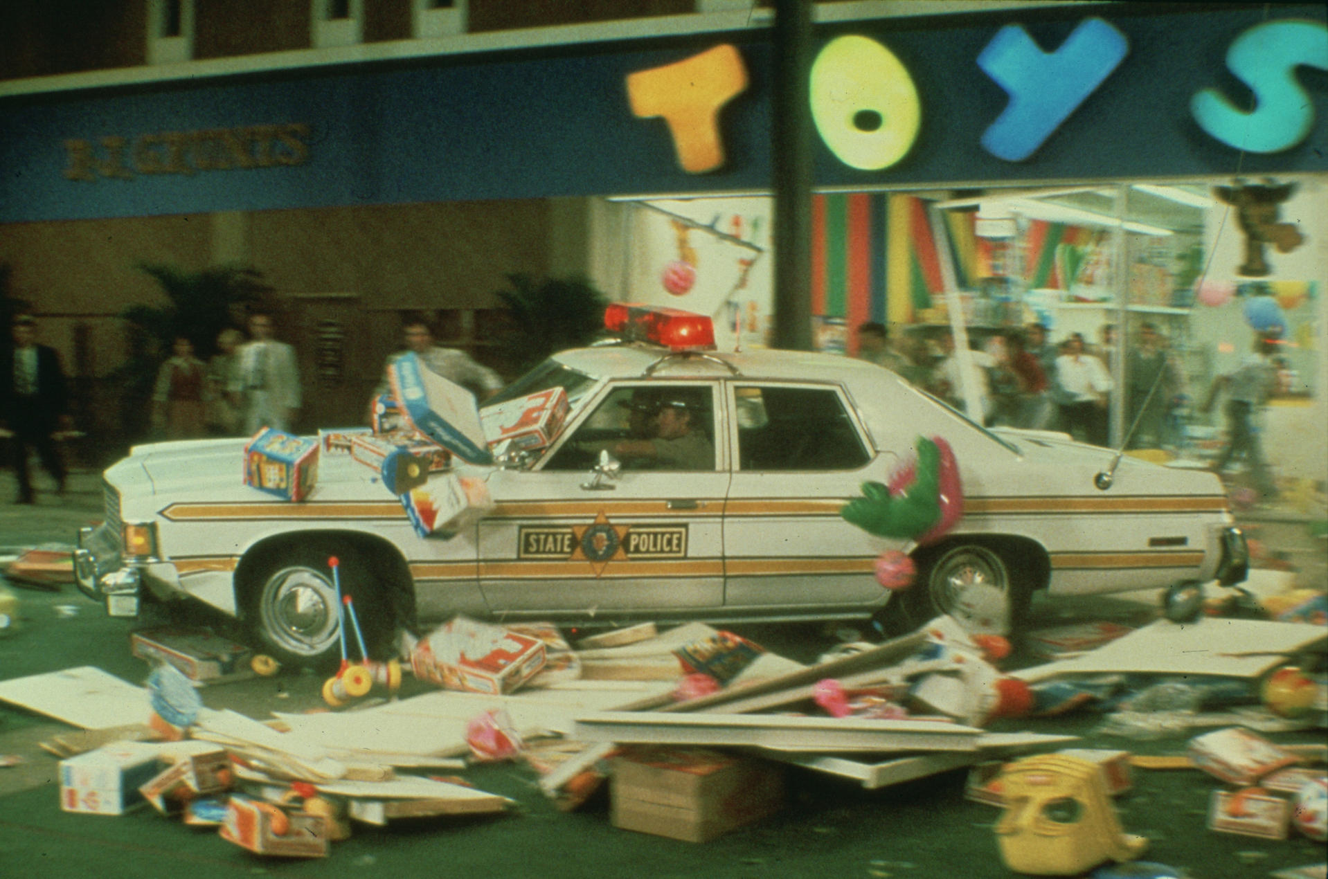 Ein weißes Polizeiauto ist von Spielzeug vor einem Toys R Us-Geschäft umgeben. Spielzeug liegt überall verstreut. Menschen sind im Hintergrund.