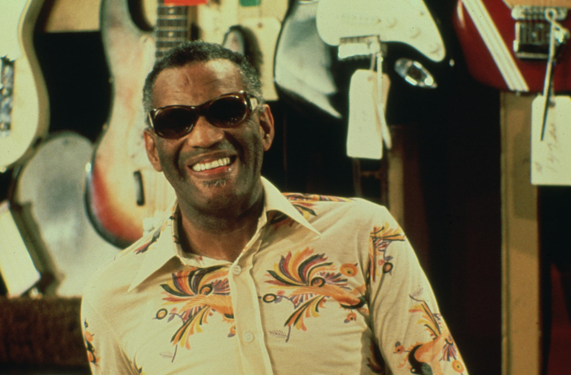 Ray Charles lächelt, trägt eine Sonnenbrille und ein gemustertes Hemd. Gitarren sind im Hintergrund.