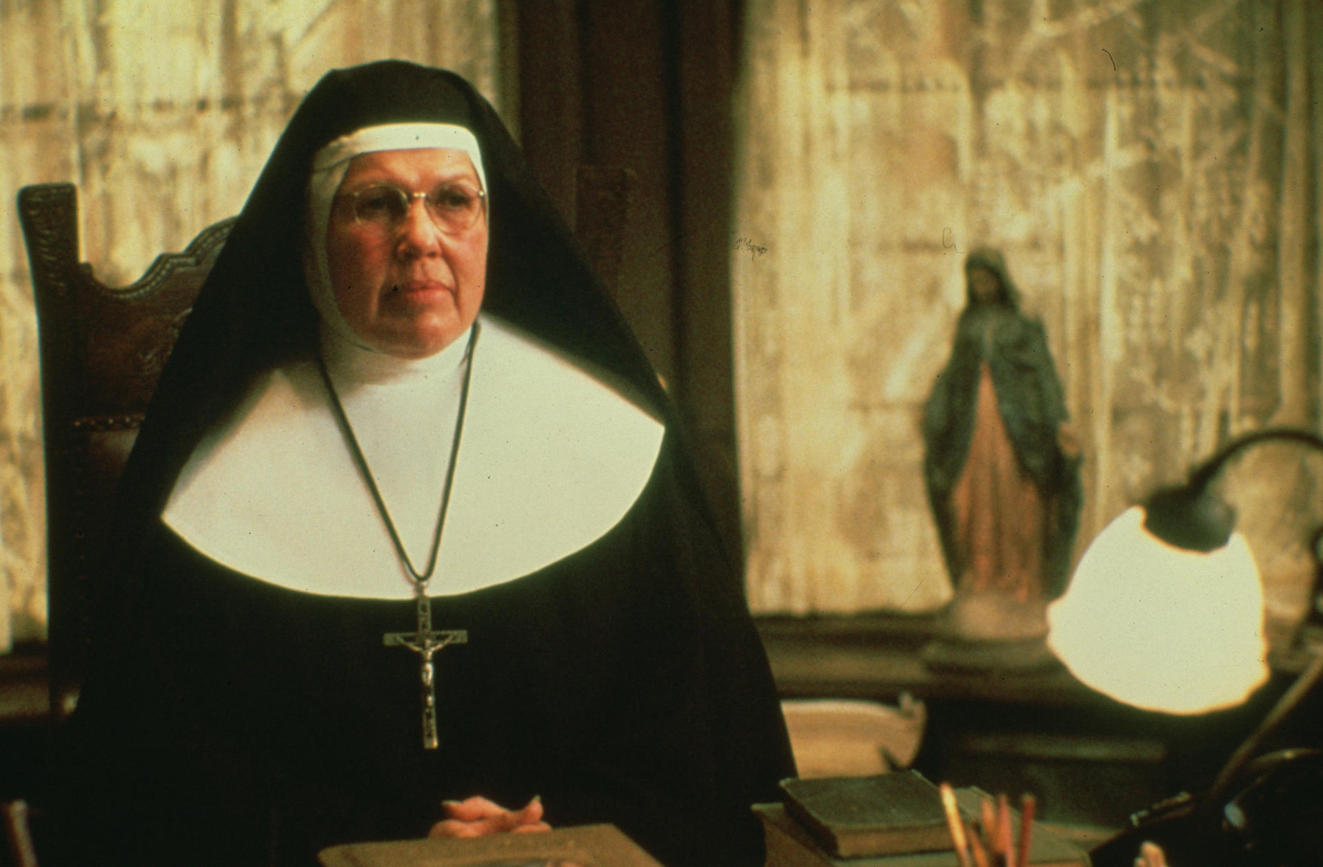 Eine Nonne in schwarzem Habit und Brille sitzt an einem Schreibtisch. Im Hintergrund eine Statue und eine Lampe. Bücher und Bleistifte liegen auf dem Tisch.