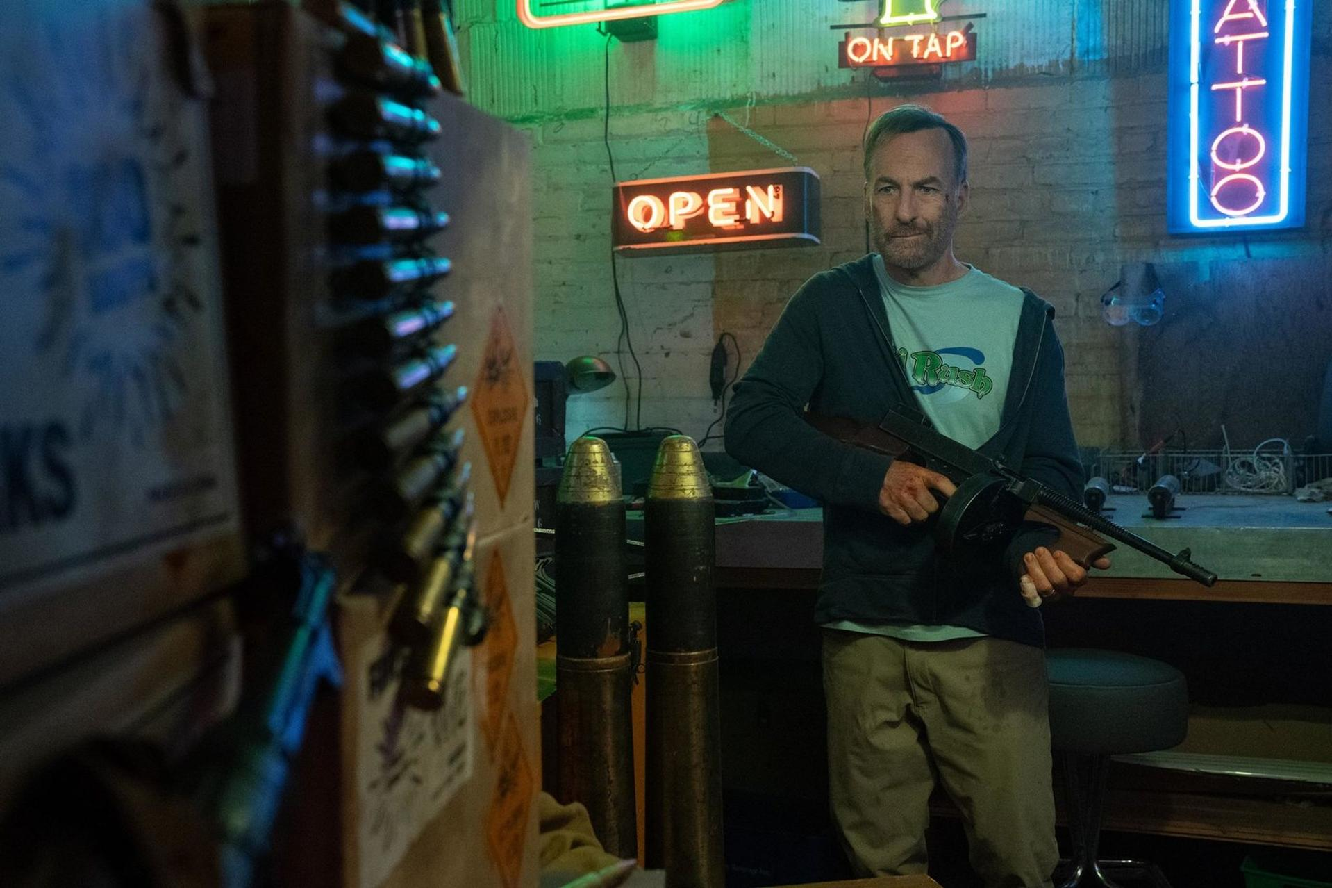 Mann mit Waffe in einem schwach beleuchteten Raum. Neon-Schilder sagen 'Open' und 'Tattoo'. Hülsen und Munitionskisten sind sichtbar.
