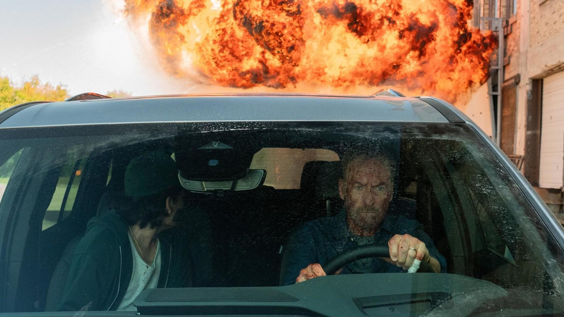 In einem Auto fährt ein Mann mit blutverschmiertem Gesicht, während draußen eine große Explosion ausbricht. Ein Passagier schaut zu.