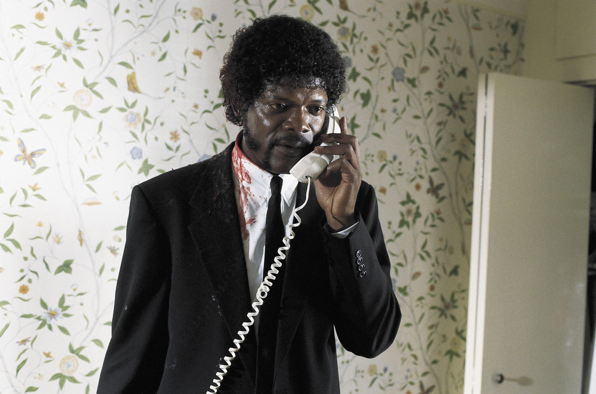 Samuel L. Jackson in einem Anzug, Blut auf dem Hemd, telefoniert. Blumige Tapete im Hintergrund.