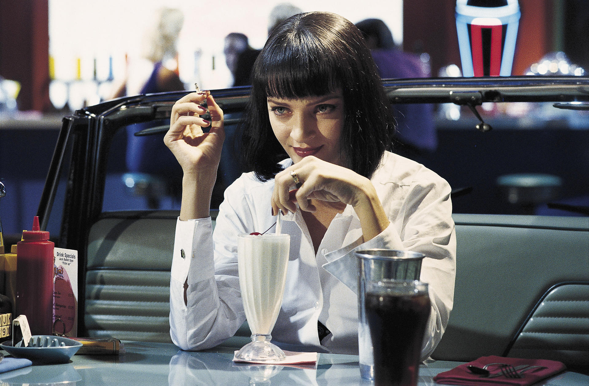 Mia Wallace sitzt rauchend an einem Tisch in einem Diner. Ein Milchshake und ein Getränk stehen auf dem Tisch. Im Hintergrund ist ein Autositz.