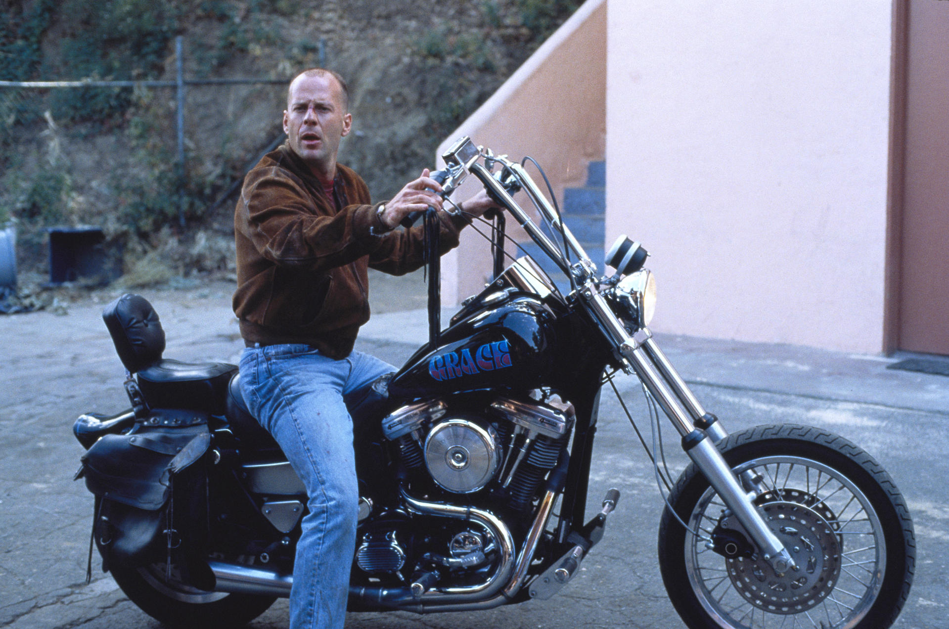Glatzkopf auf einem schwarzen Motorrad mit 'GRACE' auf dem Tank. Trägt eine braune Jacke und Jeans. Im Hintergrund ein Zaun und ein Gebäude.