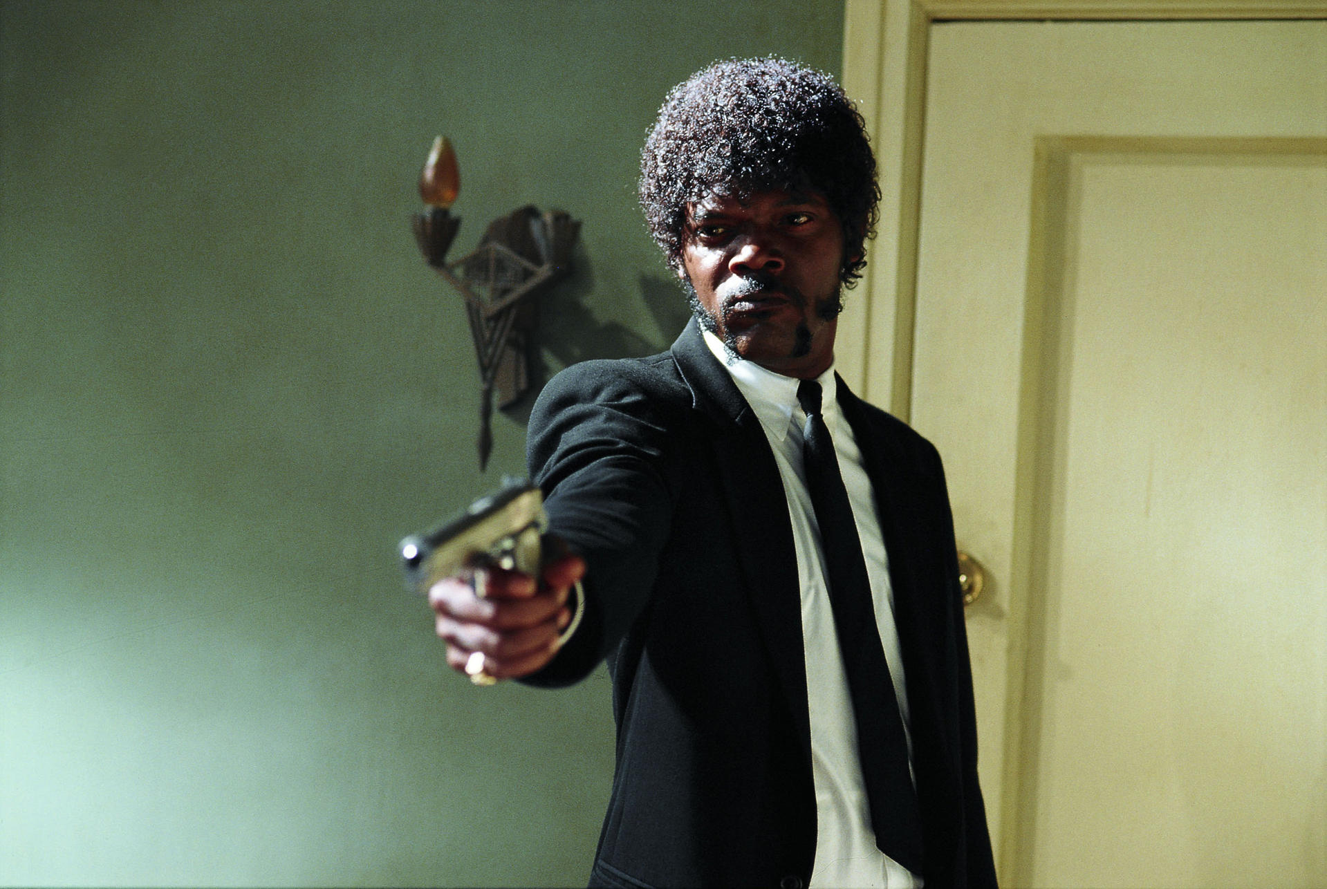 Samuel L. Jackson in einem Anzug, der eine Waffe hält. Er schaut in die Kamera. Der Hintergrund ist eine grüne Wand und eine Tür.