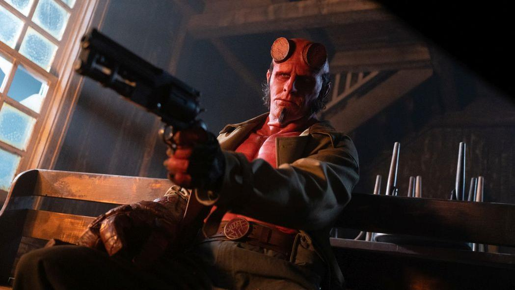 Hellboy, ein rothäutiger Dämon, sitzt und zielt mit einer Waffe. Er trägt einen beigefarbenen Mantel und hält eine Waffe. Im Hintergrund ist ein Fenster.