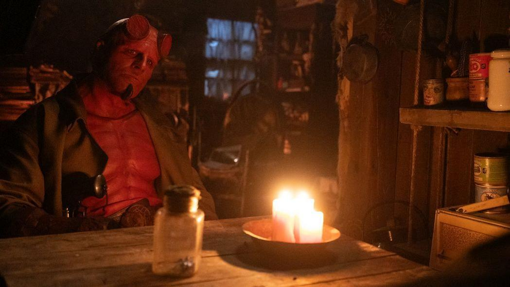 Hellboy sitzt in einem schwach beleuchteten Raum, rote Haut, Hörner und ein brauner Mantel. Kerzen auf einem Tisch davor beleuchten die Szene.