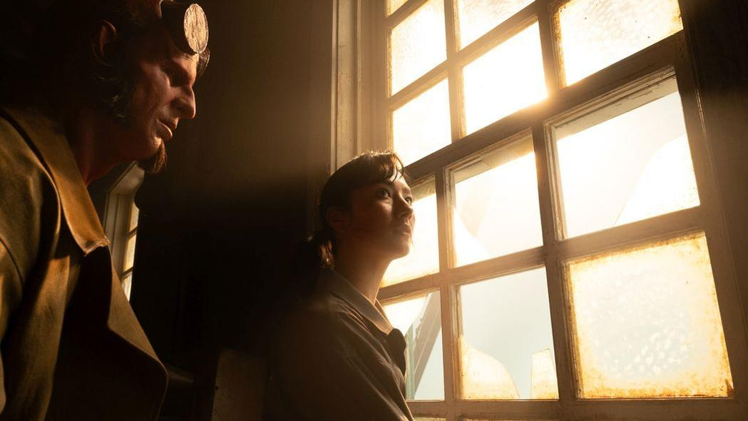 Mann mit Hörnern und Frau schauen aus einem Fenster. Sonnenlicht scheint herein und beleuchtet den Fensterrahmen und das Gesicht der Frau.