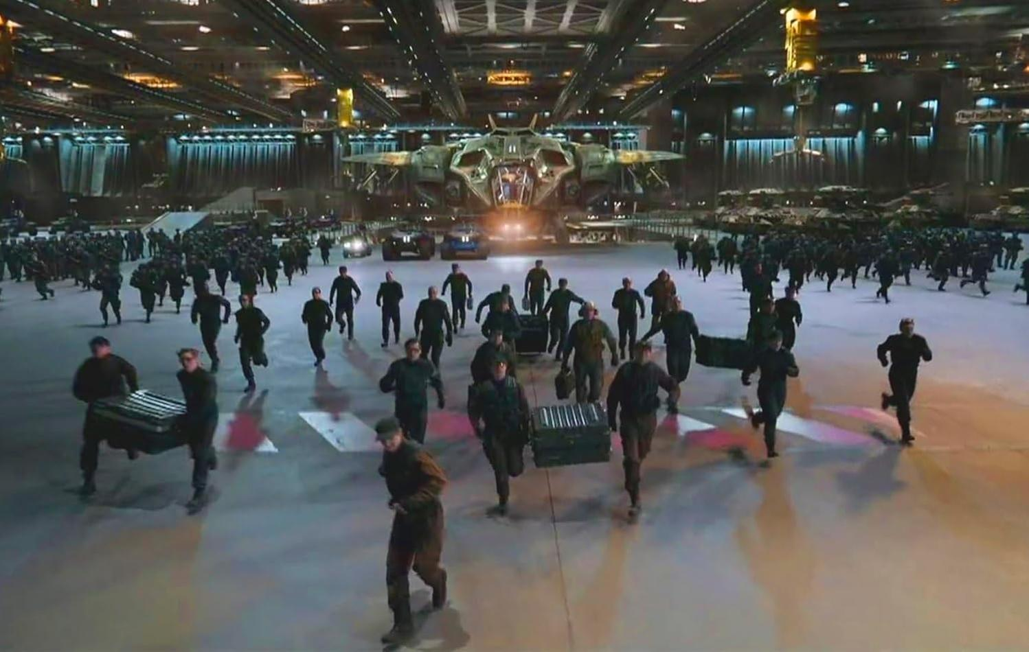 Männer rennen mit Koffern auf ein großes Flugzeug in einem Hangar zu. Im Hintergrund sind viele Fahrzeuge und Menschen zu sehen.