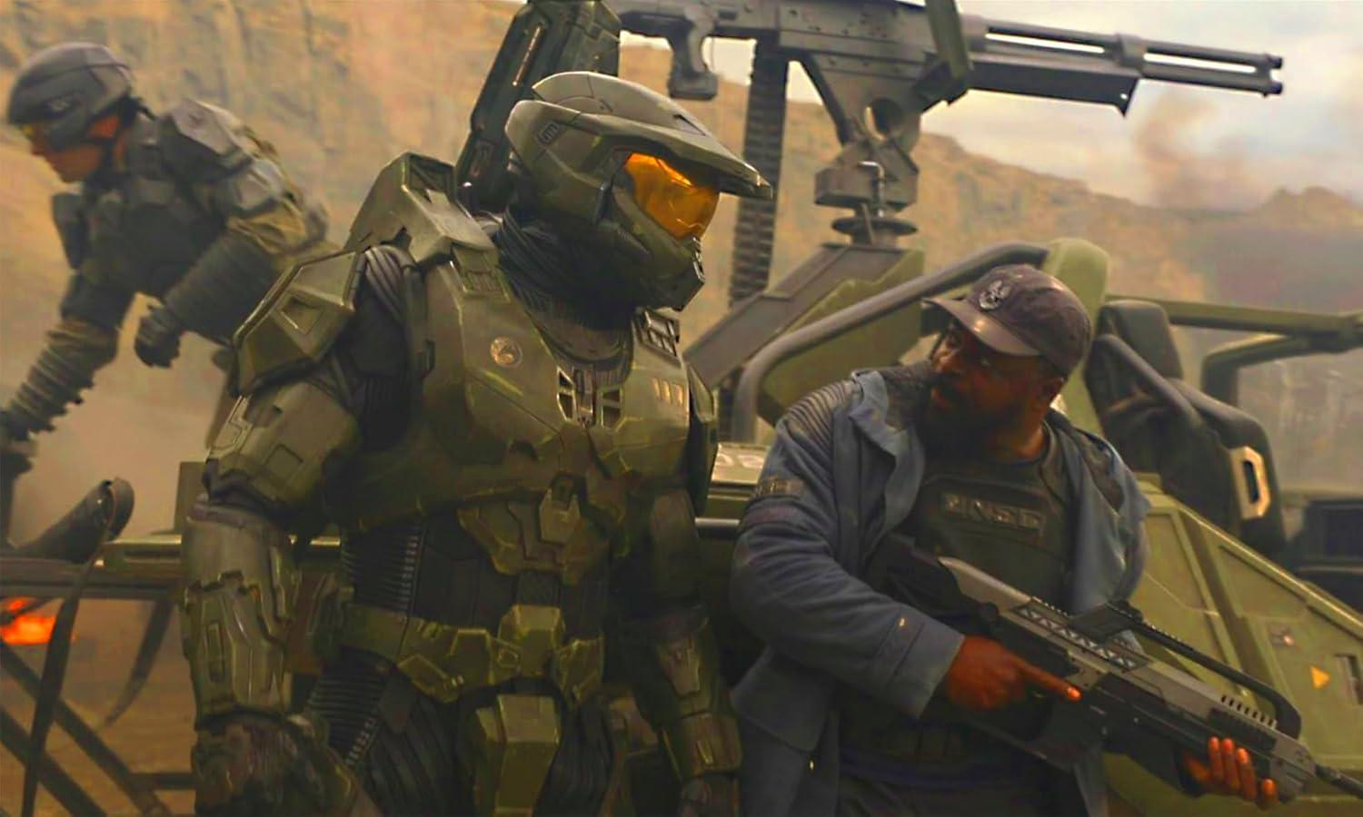 Halo-Charaktere in Rüstung und Ausrüstung stehen in der Nähe eines Fahrzeugs und einer Waffe. Eine Person im Hintergrund trägt ebenfalls Ausrüstung.