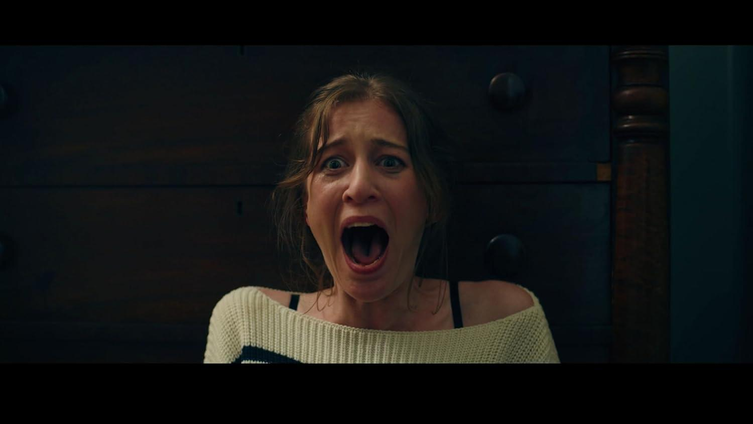 Eine Frau schreit mit aufgerissenen Augen und offenem Mund. Sie trägt einen weißen Pullover und steht vor einer dunklen Holzkommode.
