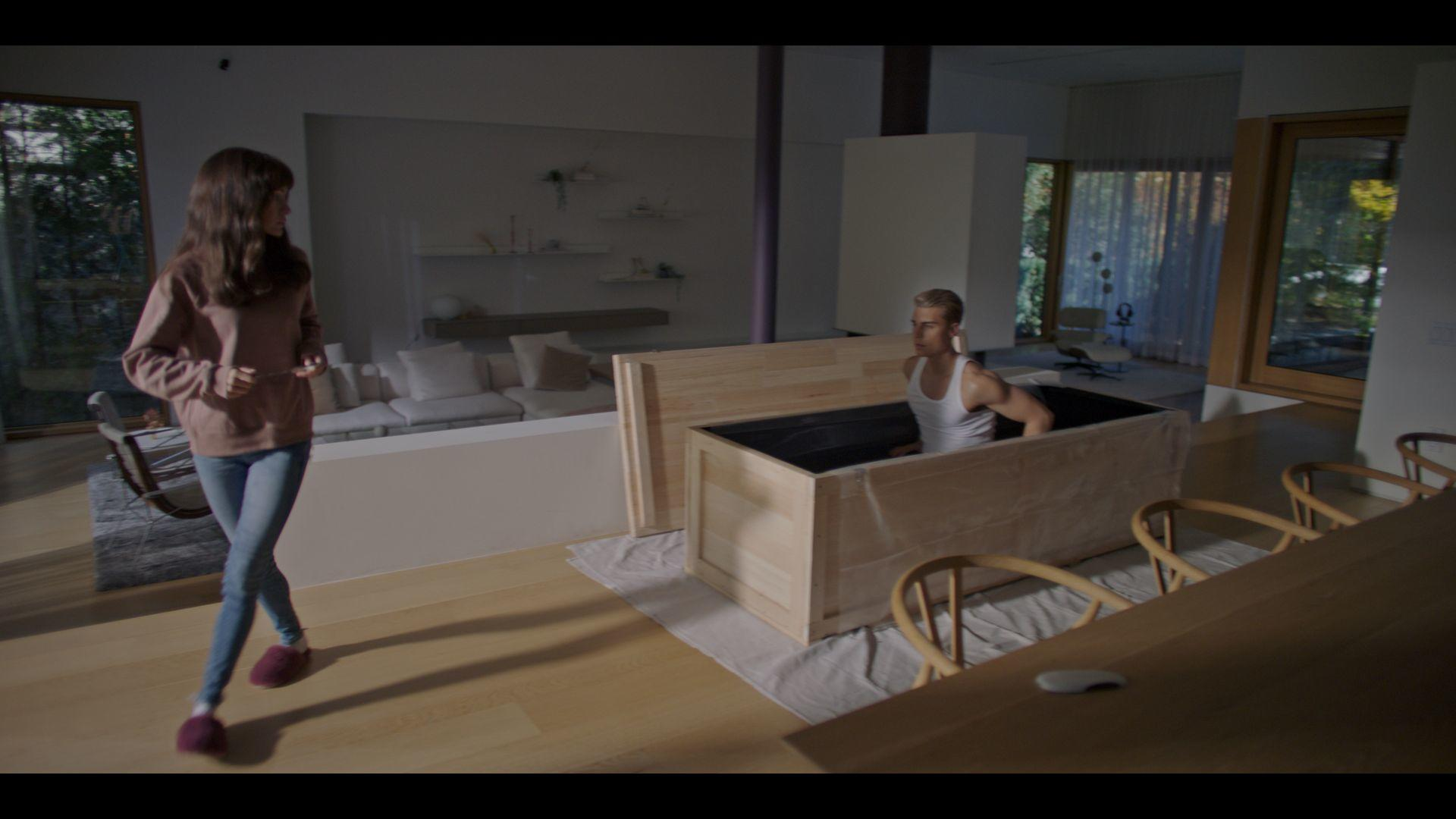 Eine Frau geht an einem Mann in einer Holzkiste vorbei. Der Raum hat große Fenster, ein Sofa und Holzstühle.