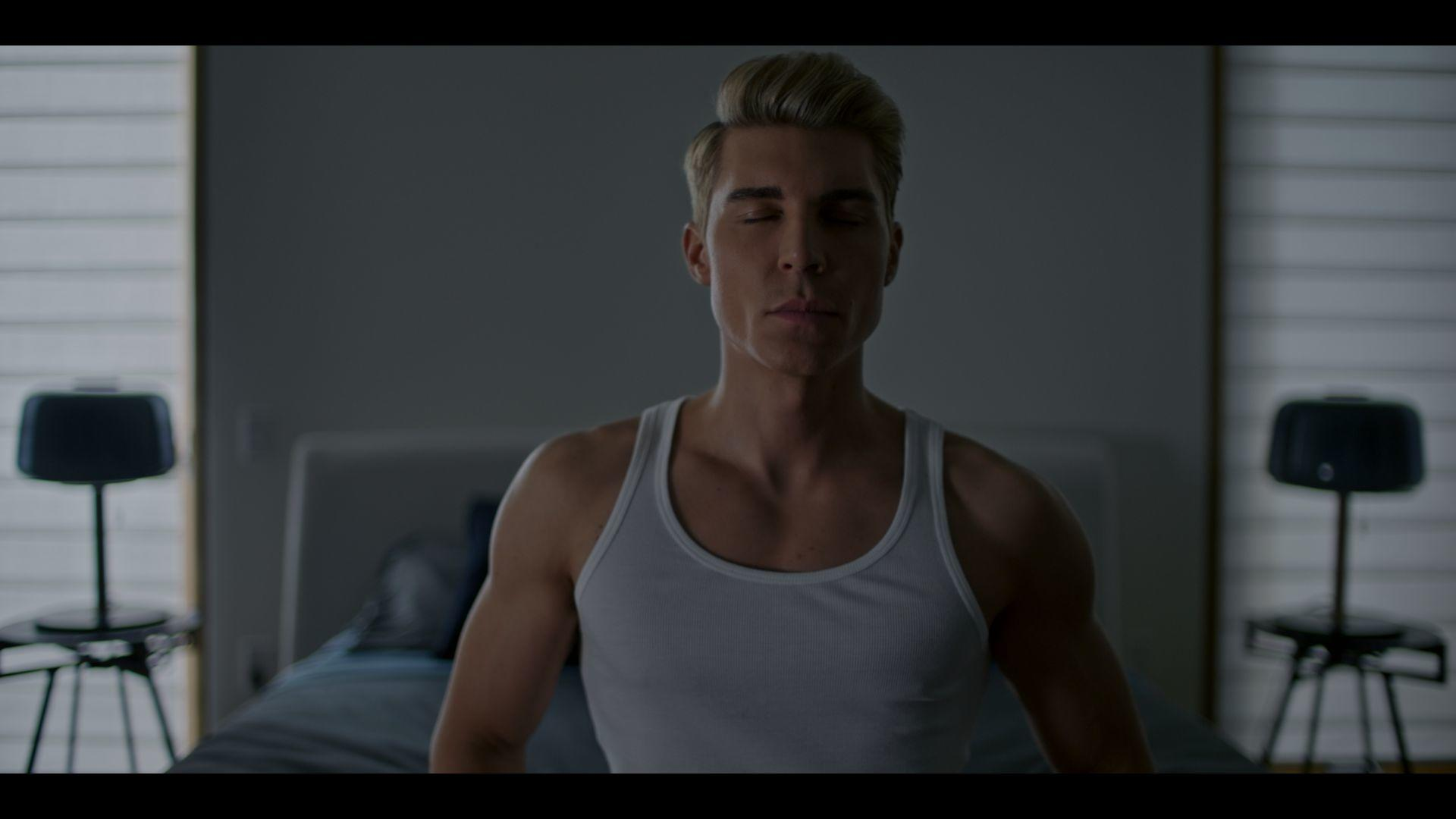 Ein Mann mit geschlossenen Augen, der ein weißes Tanktop trägt, sitzt auf einem Bett. Auf beiden Seiten stehen schwarze Stühle.