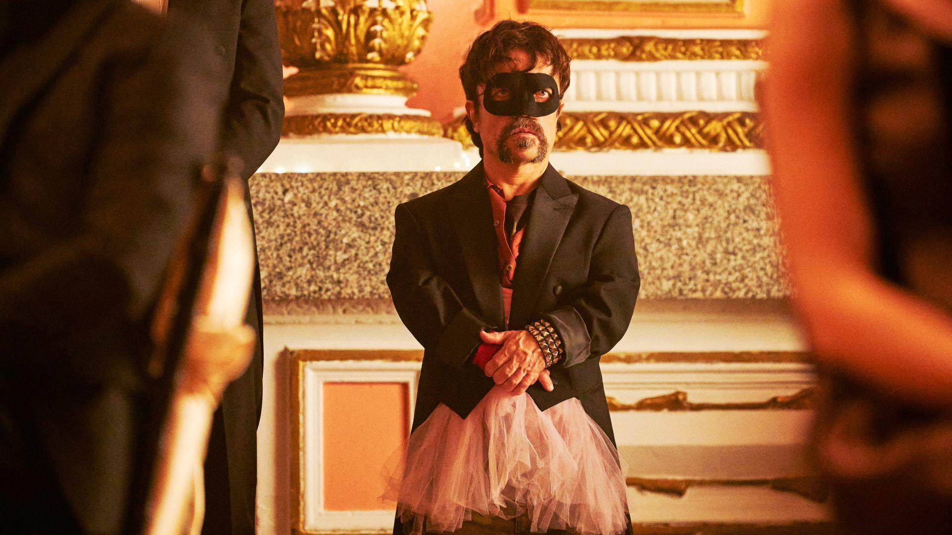 Mann mit Maske, Smoking und Tutu, steht inmitten kunstvoller Dekoration. Hände gefaltet, mit Nietenarmband.