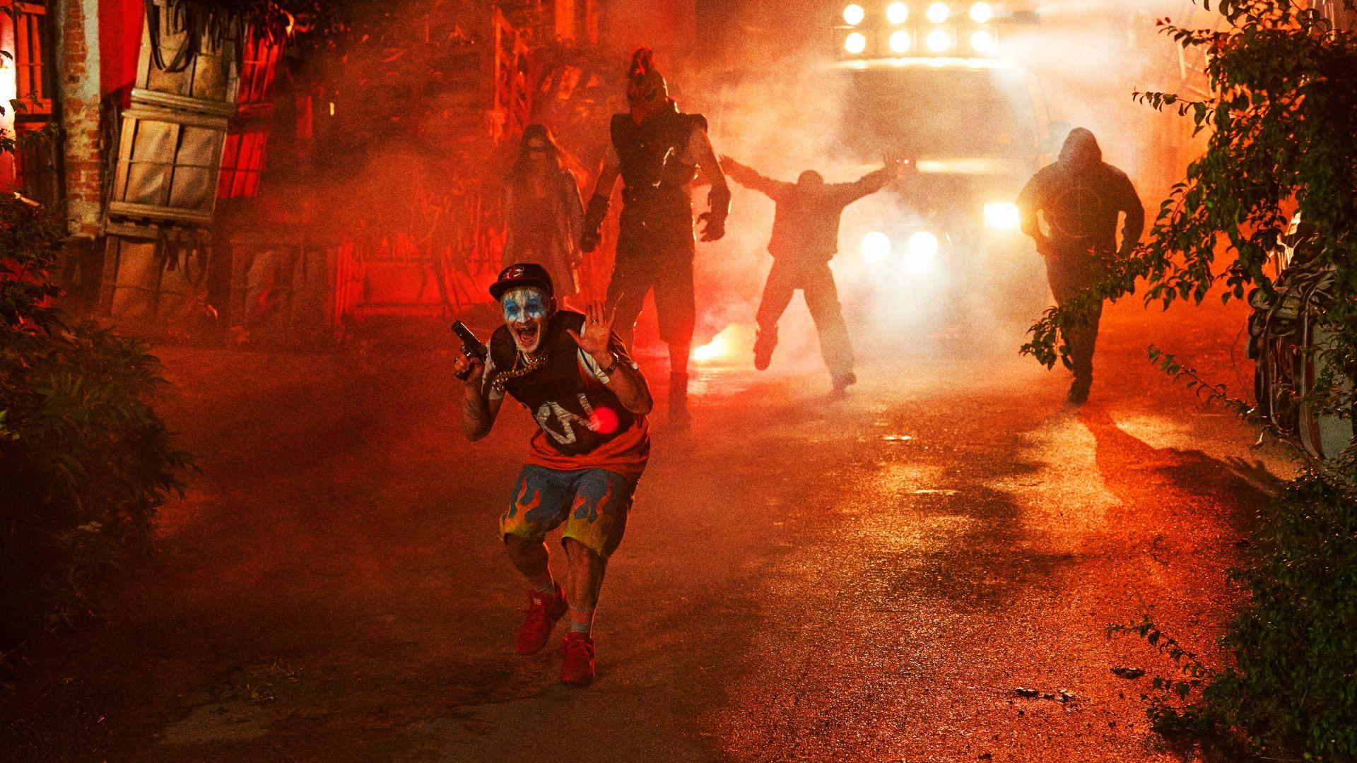 Ein Clown mit einer Waffe flieht vor Figuren im Nebel. Im Hintergrund befindet sich ein Fahrzeug mit Lichtern. Rote und orange Farbtöne dominieren.