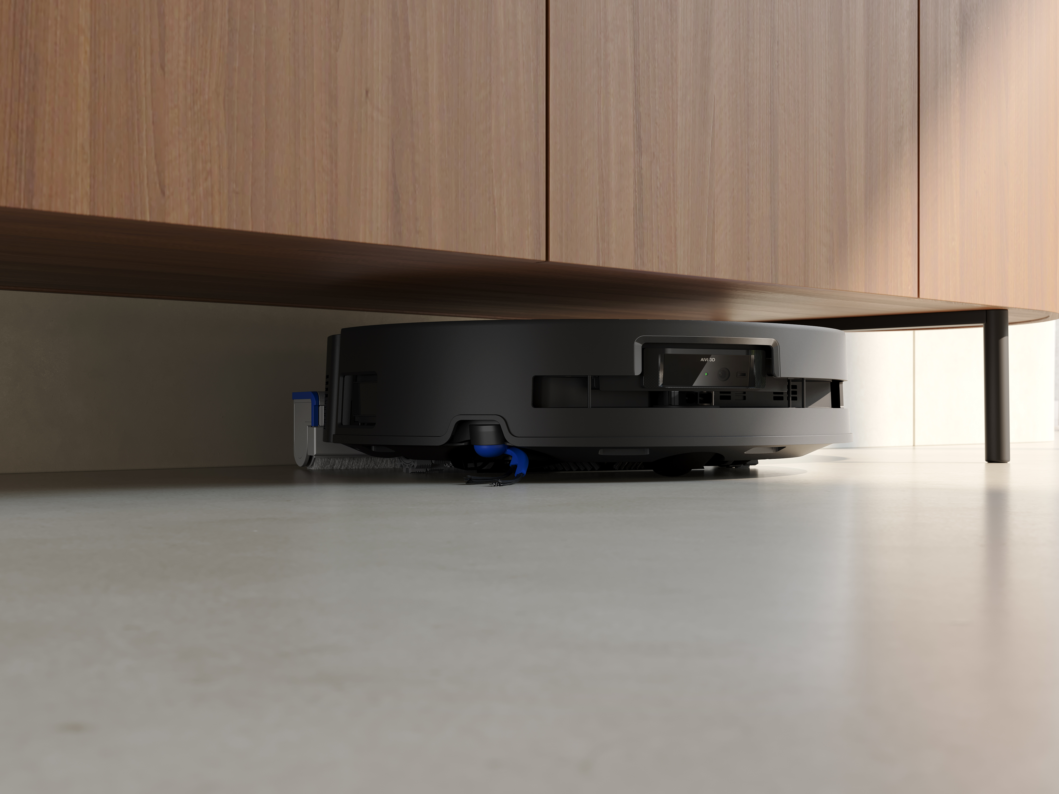 Czarny odkurzacz robot pod drewnianą szafką. Podłoga jest jasnoszara. Tło to jasne okno.