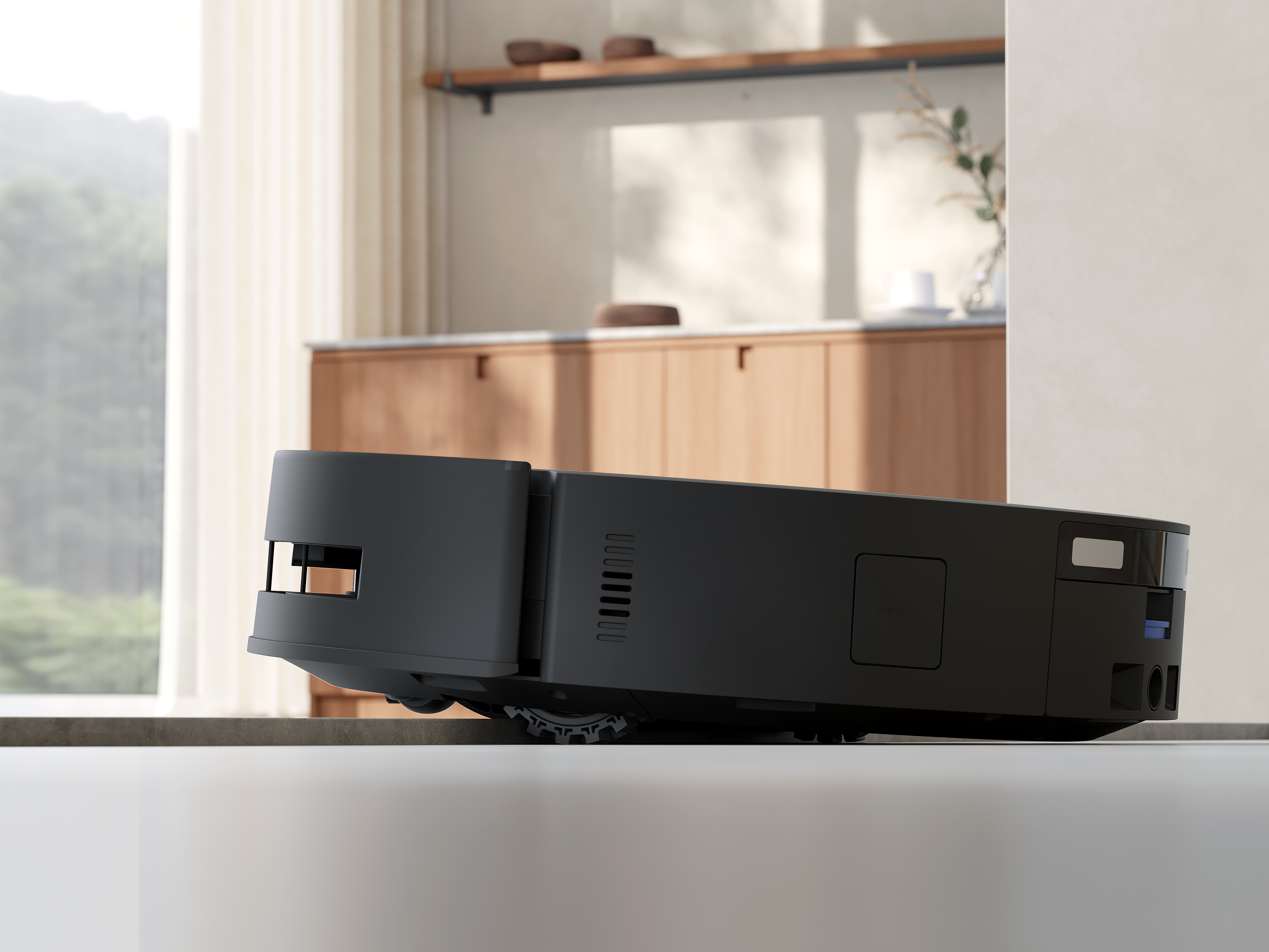 Czarny robot sprzątający na białej powierzchni. Tło obejmuje okno i drewniane meble.