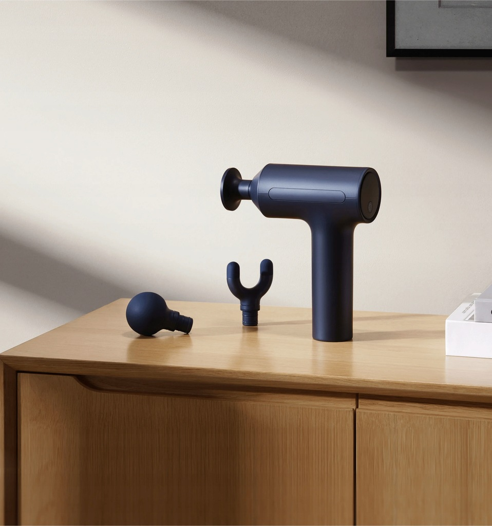 Granatowy pistolet do masażu z trzema wymiennymi końcówkami na drewnianej szafce. Biała ściana i oprawiona grafika w tle.