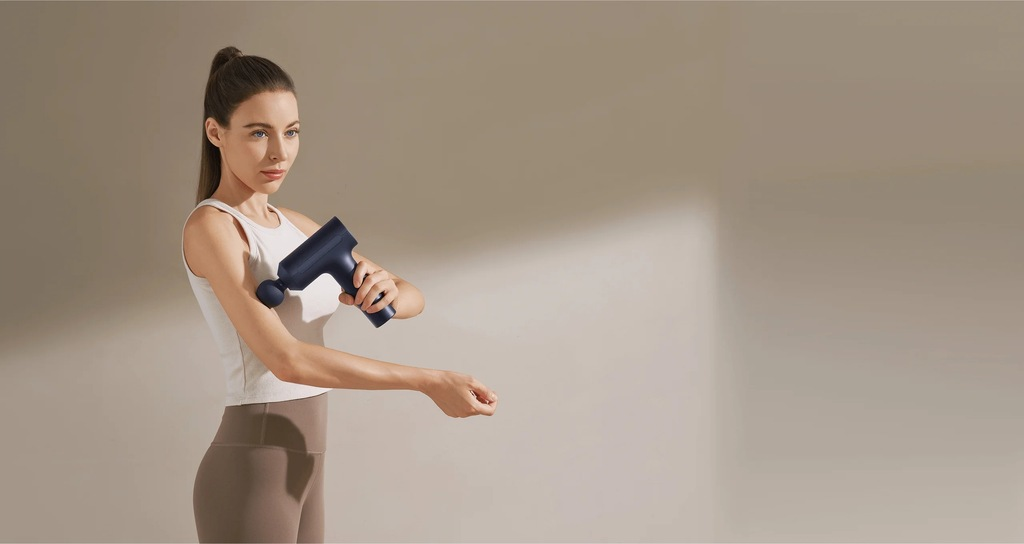 Kobieta w stroju do ćwiczeń używa pistoletu do masażu na ramieniu. Tło jest jednolite, beżowe.