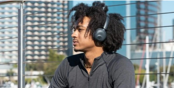 Mężczyzna ze słuchawkami patrzy w lewo. Ma na sobie szarą koszulę i kręcone włosy. W tle budynki i balustrada.