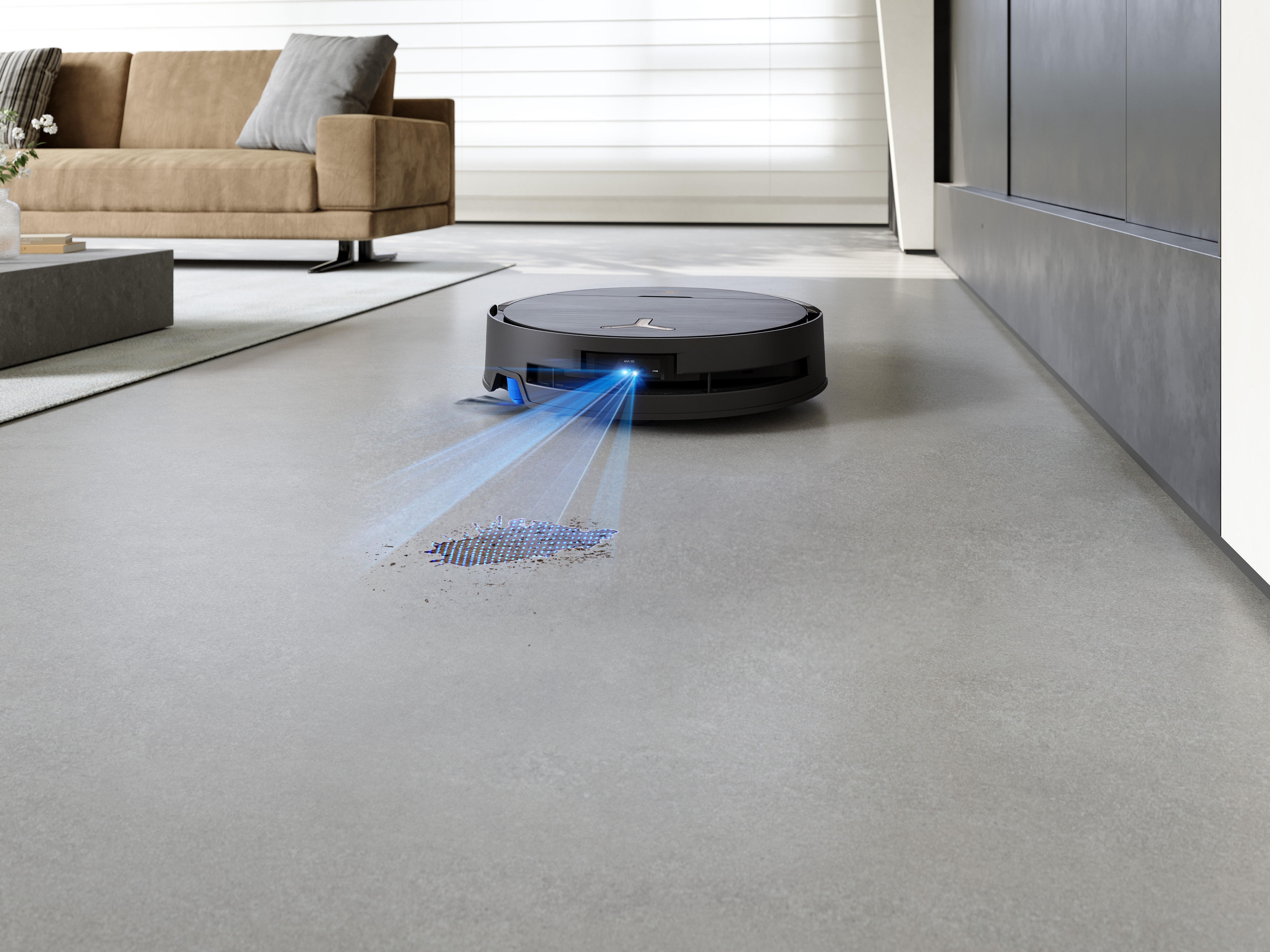 Robot sprzątający na szarej podłodze, w trakcie sprzątania. Brązowa sofa i szafka w tle.
